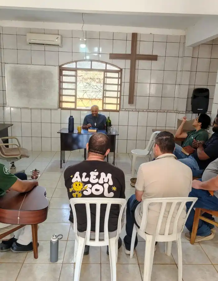 Grupo de homens reunido em círculo para estudo bíblico no salão do campus, com uma cruz de madeira na parede