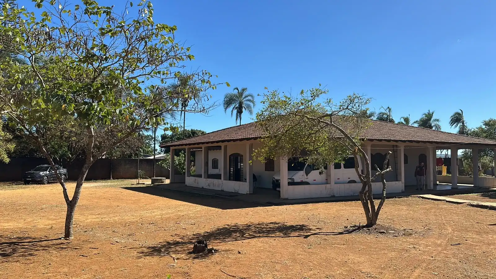 Casa principal do campus com telhado de cerâmica, varanda ampla, árvores e palmeiras sob céu azul