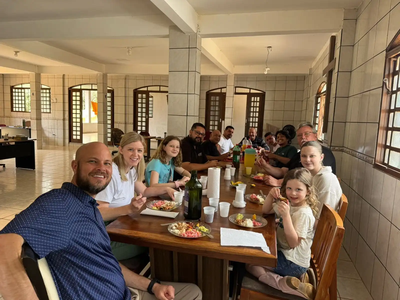 Participantes, equipe e família visitante almoçando juntos em uma longa mesa de madeira no refeitório