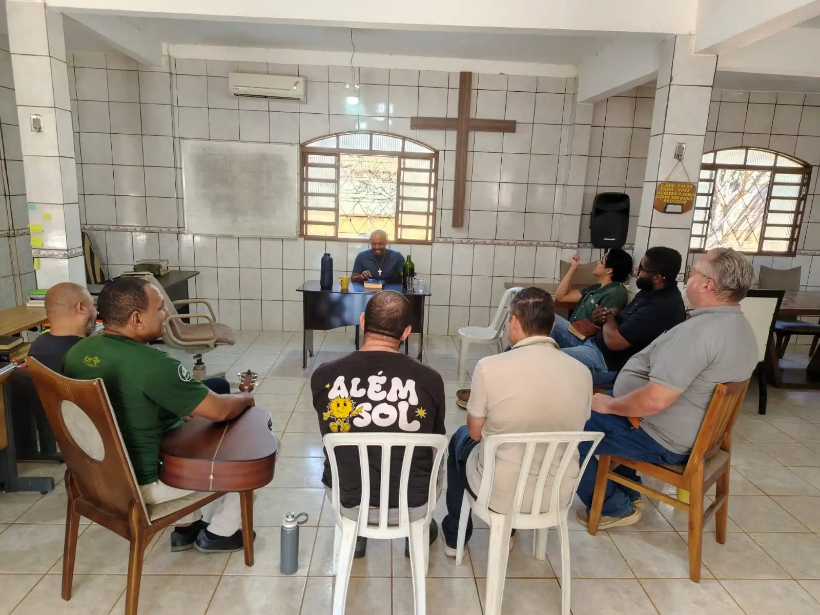 Homens sentados em círculo durante estudo bíblico no salão principal, com uma grande cruz de madeira na parede